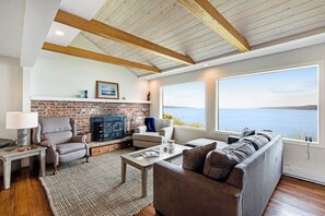 Ein Wohnzimmer mit Kamin, einer Couch, einem Couchtisch und großen Fenstern mit Blick auf ein Gewässer.