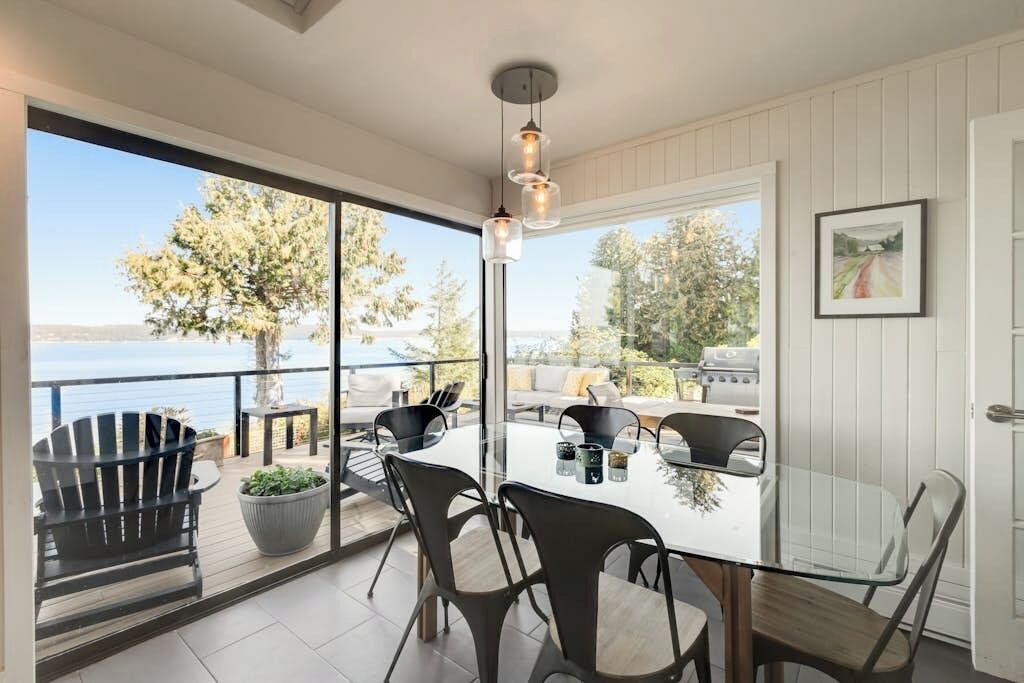 A dining area with a glass table and chairs, a view of the sea, and a barbecue grill outside.