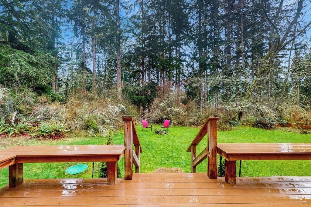 A wooden deck with a view of a forested area, a grassy lawn, and two pink chairs.