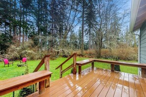 A wooden deck with a wooden railing, a grassy yard, and a forest in the background.