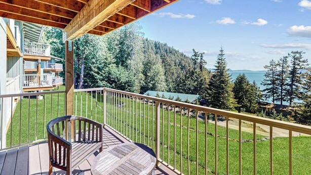 A wooden deck with a railing, a chair, and a view of a forest and lake. - Wonderful Waterfront Escape with Three Balconies and Gas Fireplace (Eastsound)