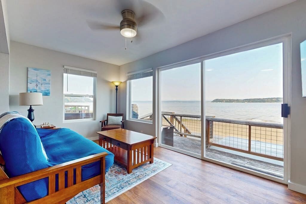 Ein Zimmer mit Bett, Tisch und Blick auf das Wasser durch große Fenster.