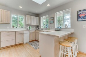 A kitchen with light wood cabinets, a central island with stools, and a window overlooking greenery. - Stunning Waterfront Home on Lopez Island with Seasonal Hot Tub (Lopez Island)