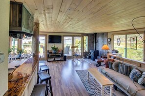 Uma sala de estar ampla com teto de madeira, uma mesa de jantar comprida, um sofá e uma televisão.