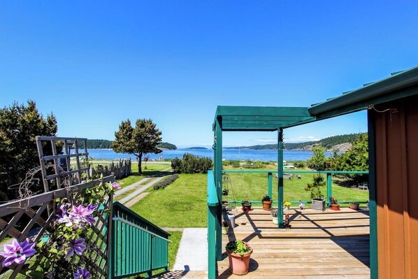 Eine Holzterrasse mit Blick auf einen See und Hügel, eine grüne Pergola und Kübelpflanzen.