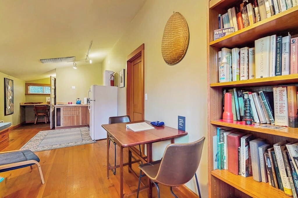 A room with a wooden dining table, chairs, a bookshelf, a kitchen area, and a large woven wall hanging.