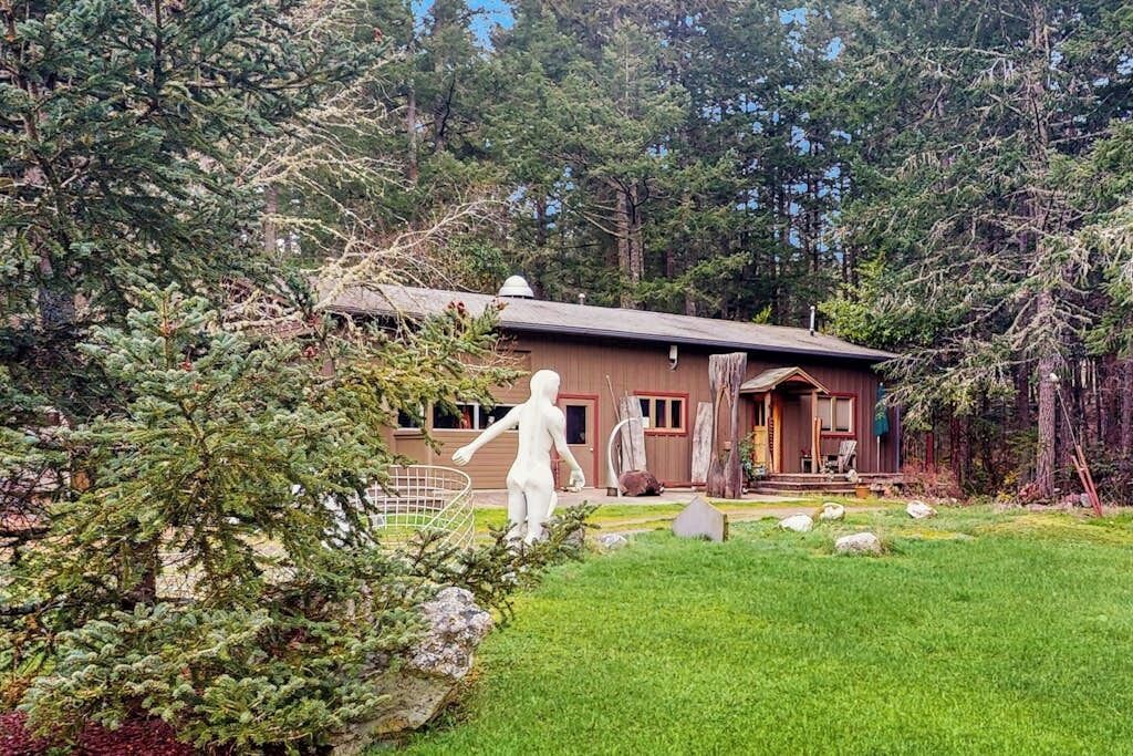 Une cabane aux murs en bois, entourée d’une zone boisée avec une pelouse verdoyante et quelques rochers.