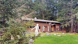 A cabin with a wooden exterior, surrounded by a forested area with a grassy lawn and a few rocks. - Dog Friendly Studio with Kitchenette, WiFi, Yard and Fire Pit near Eastsound Beach (Eastsound)