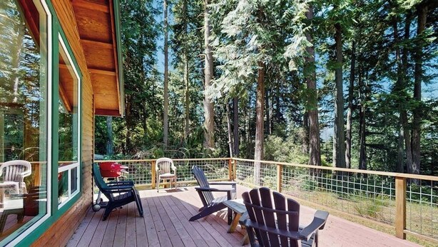 Eine Holzterrasse mit Adirondack-Stühlen und einem Tisch, mit Blick auf ein bewaldetes Gebiet.