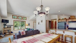 Ein geräumiger Wohnbereich mit einer großen Couch, einem Esstisch mit Bänken, einer Küche mit Kühlschrank und einer Kristallleuchtre.