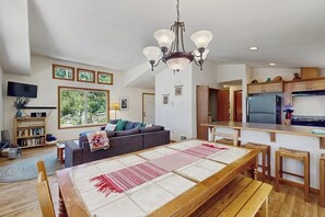 Een ruime woonkamer met een grote bank, een eettafel met banken, een keuken met een koelkast, en een kroonluchter.