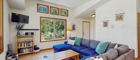 Una sala de estar con un sofá modular, una mesa de centro, una estantería, un televisor de pantalla plana y un ventanal con vistas a zonas verdes.