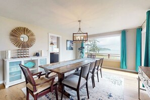 A dining area with a long wooden table, chairs, a chandelier, and a view through large windows.