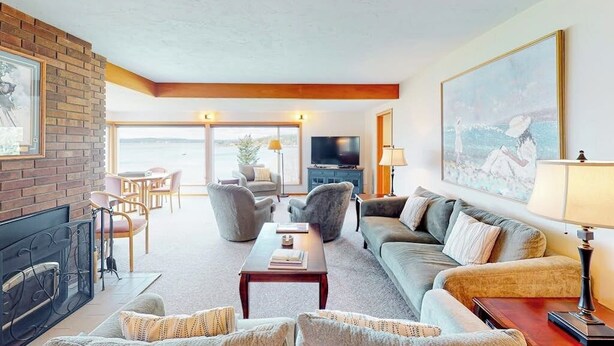 A living room with a fireplace, sofa, and television. There is a large window with a view. - Stunning Waterfront Home on Fisherman Bay with Sunset Views and Ping Pong (Lopez Island)