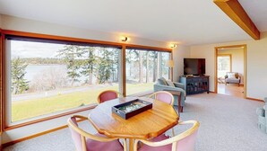 A spacious room with a large window, a dining table with chairs, a sofa, and a television. - Stunning Waterfront Home on Fisherman Bay with Sunset Views and Ping Pong (Lopez Island)