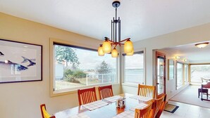 A dining area with a wooden table, chairs, and a chandelier. There are large windows providing a view of the outdoors. - Stunning Waterfront Home on Fisherman Bay with Sunset Views and Ping Pong (Lopez Island)