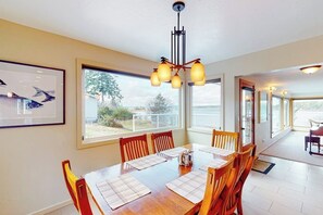 A dining area with a wooden table, chairs, and a chandelier. There are large windows providing a view of the outdoors.