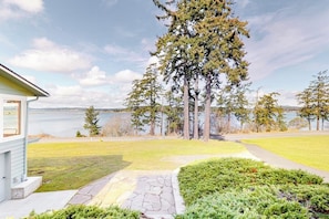 A residential property with a stone pathway leading to a grassy area, a small house, and a view of a lake with trees.