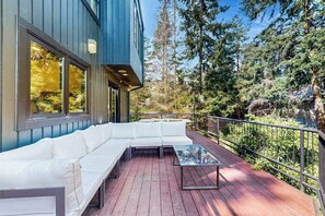 Une terrasse extérieure moderne, avec un canapé blanc et une table en verre, entourée d’une rambarde en métal et d’une forêt dense.