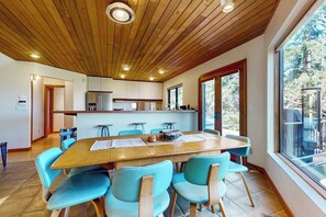 Une cuisine moderne avec un plafond en bois, un coin repas doté d’une grande table et de chaises bleues, et une vue sur l’extérieur grâce à de grandes fenêtres.