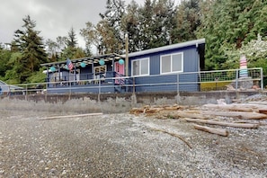 A building with a covered deck and a flag, surrounded by trees and a body of water. - Stunning Soundfront Home in Coupeville with Private Beach Access and Fire Pit (Coupeville)