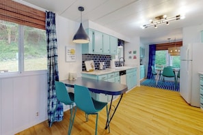 A modern kitchen with a dining area, featuring light blue cabinets, a wooden floor, and a view of the outdoors. - Stunning Soundfront Home in Coupeville with Private Beach Access and Fire Pit (Coupeville)