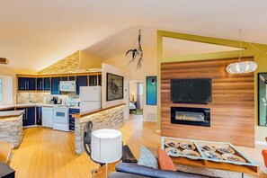 A modern kitchen with a stone backsplash, a dining area with a wooden table, and a living room with a fireplace. - Unique Treetop Home with 360 Degree Deck (Eastsound)