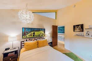 A bedroom with a bed, bedside tables, a chandelier, artwork, and a view to a balcony. - Unique Treetop Home with 360 Degree Deck (Eastsound)