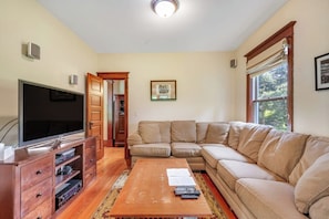 Una sala de estar con una televisión, una mesa de centro de madera, un sofá color beige y una puerta que da a otra habitación.