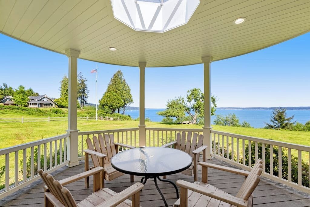 A covered outdoor seating area with a round table and chairs, overlooking a body of water and a grassy landscape.