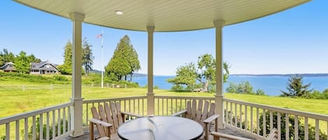 A covered outdoor seating area with a round table and chairs, overlooking a body of water and a grassy landscape.