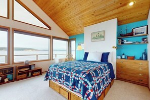 A bedroom with a large bed, wooden ceiling, a view through the windows, and a shelf with decorative items. - Relaxing Beachfront Retreat with Panoramic Water Views and Spacious Outdoor Deck (Langley)
