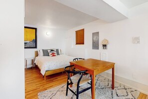 Een slaapkamer met een bed, een houten tafel, een stoel, een klein nachtkastje, een wandklok en een lijst met een afbeelding.