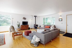Een moderne woonkamer met een grijze bank, houten vloer, een televisie, een open haard en grote ramen.