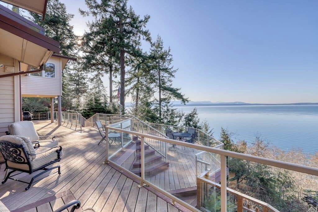 A wooden deck with outdoor furniture, a glass railing, and a view of a lake and mountains.