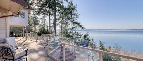 A wooden deck with outdoor furniture, a glass railing, and a view of a lake and mountains.