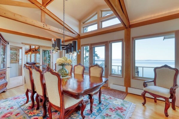 A dining room with a wooden table and chairs, large windows, and a view of the outdoors.