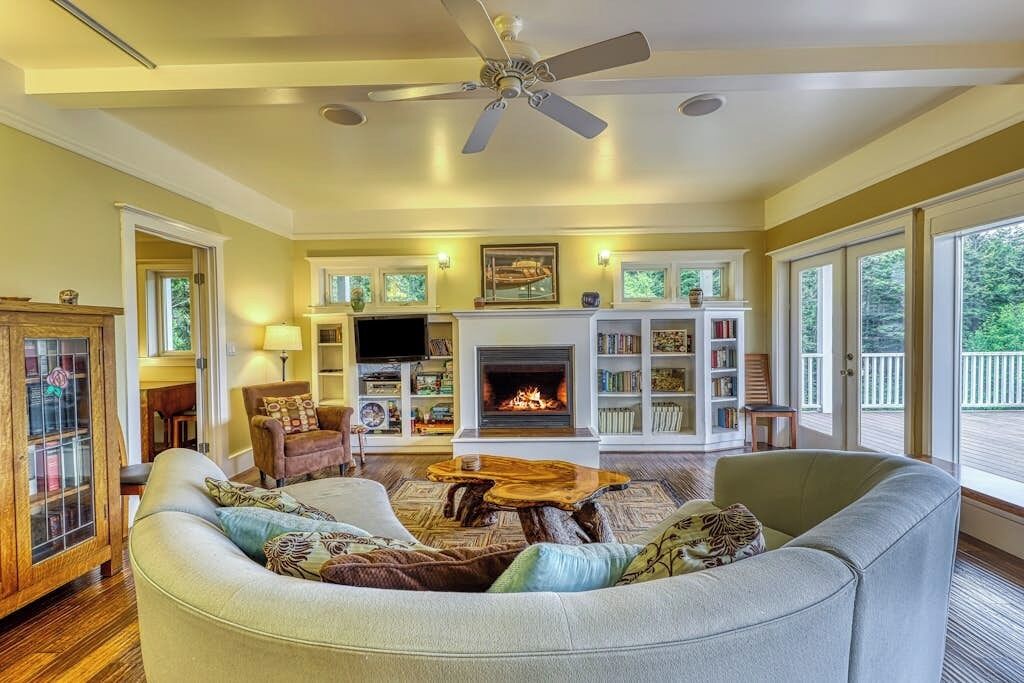A living room with a fireplace, bookshelves, a ceiling fan, and a large window.