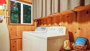 A laundry area with a washing machine, a window with curtains, and a basket with a book. - Quiet 2 BR Lopez Island Cabin Near Beach (Lopez Island)