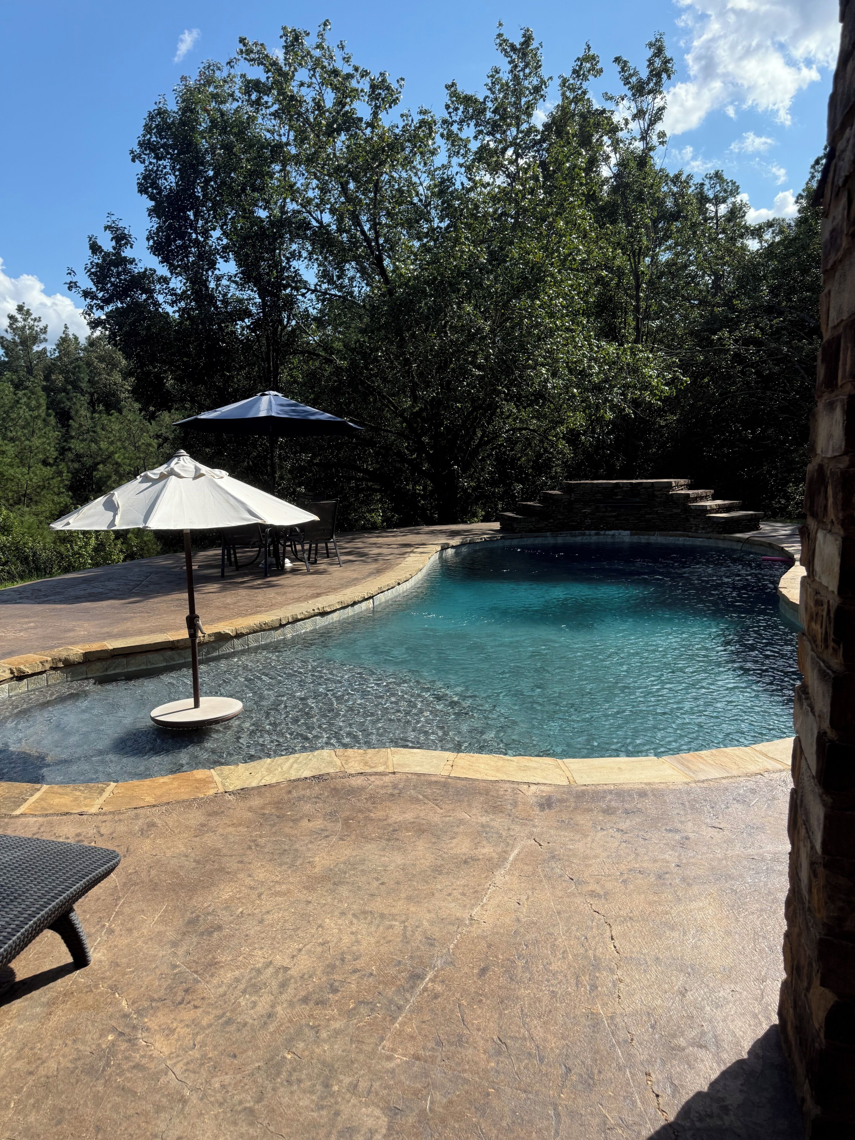 Una piscina con borde de piedra, rodeada por un patio con sillas y sombrillas.