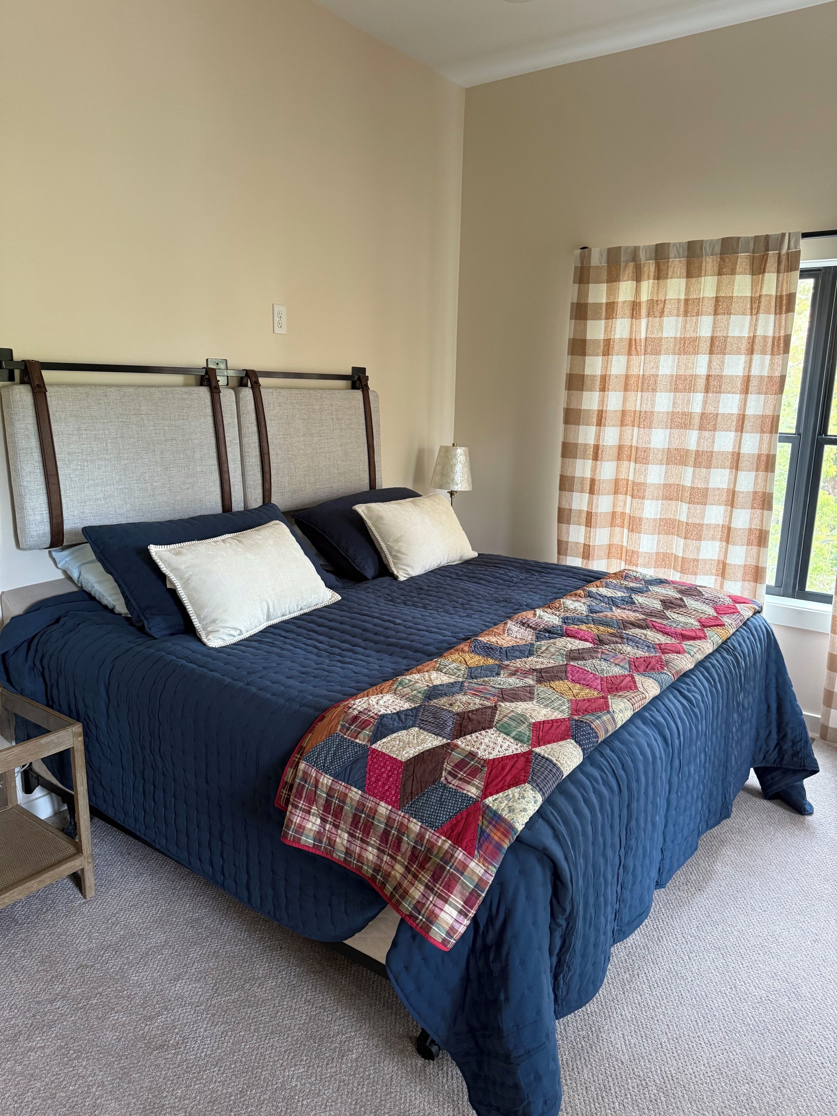 A bedroom with a bed, pillows, a quilt, a nightstand, and a window with curtains.