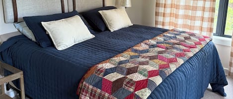 A bedroom with a bed, pillows, a quilt, a nightstand, and a window with curtains.