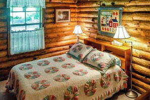 Una camera in una baita di legno, con un letto, due lampade, una coperta trapuntata, una finestra con tende e un elemento decorativo a parete.