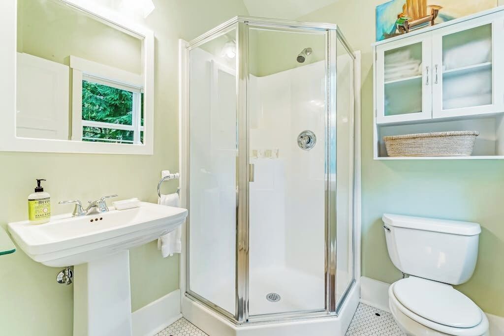 Un bagno con doccia vetro, lavabo, water e mobile contenitore con cesto.
