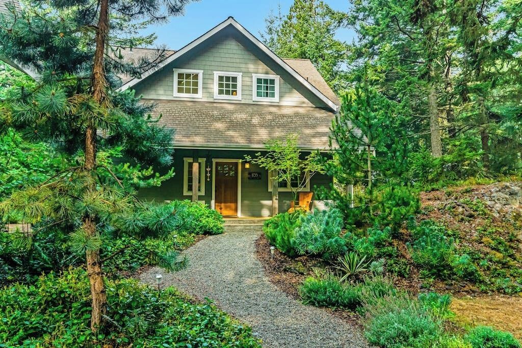 A two-story house with a front porch, surrounded by dense greenery and a gravel pathway leading to the entrance.