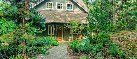 A two-story house with a front porch, surrounded by dense greenery and a gravel pathway leading to the entrance.