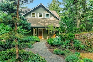 A two-story house with a front porch, surrounded by dense greenery and a gravel pathway leading to the entrance.