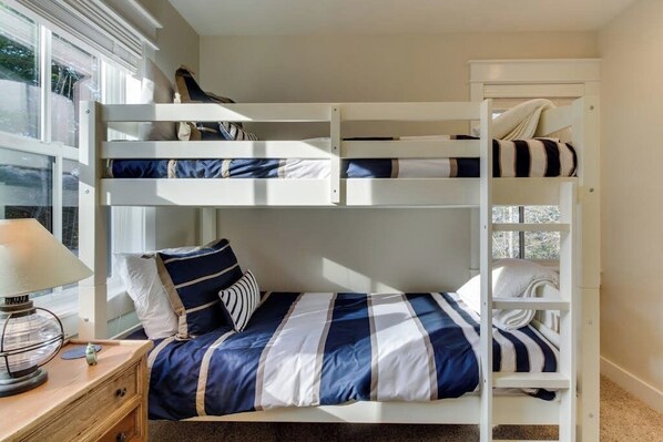 A bunk bed with a striped bedspread, a bedside table with a lamp, and a window with blinds.