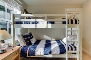 A bunk bed with a striped bedspread, a bedside table with a lamp, and a window with blinds.