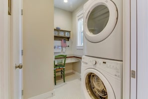 A laundry room with a stacked washer and dryer, a chair, and a shelf with items.